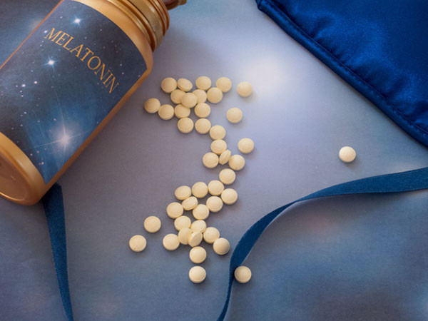 overhead view of a bottle of melatonin supplements on its side lying next to a pile of pills and a sleep mask
